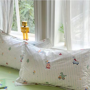 Puede incluir: Dos fundas de almohada blancas con rayas verticales azul claro y un patrón repetitivo de dinosaurios coloridos sobre monopatines y patinetes, intercalados con pequeños árboles, estrellas y globos, con un robot amarillo de fondo.