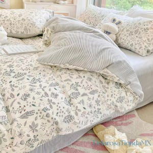 Puede incluir: Una cama con una funda nórdica y fundas de almohada con estampado floral. La funda nórdica tiene un fondo blanco con diseños botánicos grises y verdes. Un edredón a rayas grises y blancas está doblado sobre la funda nórdica. Un conejito de peluche blanco está en la cama.