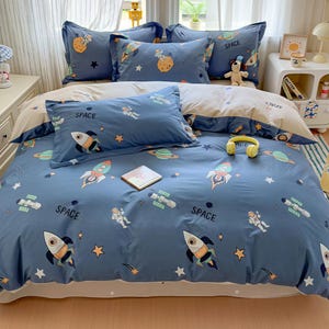 Puede incluir: Juego de cama azul con temática espacial que presenta cohetes, planetas, astronautas y estrellas. El juego incluye una funda nórdica y varias fundas de almohada. La palabra "SPACE" está impresa en la ropa de cama.