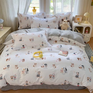 Puede incluir: Juego de cama blanco con un divertido estampado de perros. El juego incluye una funda nórdica y fundas de almohada, todas adornadas con ilustraciones de perros. La funda nórdica tiene un borde a cuadros blanco y negro. Un peluche de perro está en la cama.