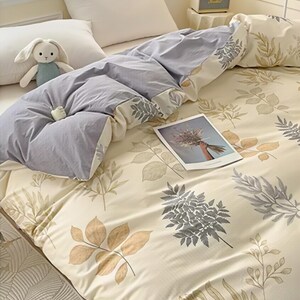 Puede incluir: Una cama con un edredón azul claro y un juego de sábanas color crema. Las sábanas tienen un estampado de hojas en tonos grises y marrones. Un pequeño conejito de peluche y un marco de fotos también están en la cama.