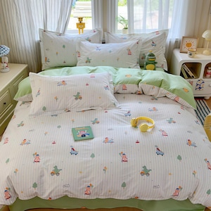 Puede incluir: Un juego de cama blanco y verde con un patrón caprichoso de personajes de dibujos animados, vehículos y árboles. El juego incluye una funda nórdica, fundas de almohada y una sábana bajera. Un libro pequeño y unos auriculares amarillos están en la cama.