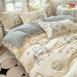 Puede incluir: Juego de cama color crema con un divertido estampado de osos y conejos en azul marino. El juego incluye fundas de almohada, una funda nórdica y una manta a cuadros. El diseño presenta osos de dibujos animados, conejos e ilustraciones de cajas de regalo.