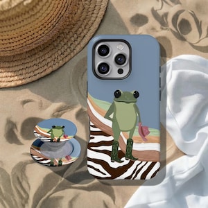 Puede incluir: Una funda de smartphone azul y un soporte circular a juego con una rana verde que lleva botas verdes y sostiene un sombrero rosa. El diseño incluye un fondo de rayas onduladas en tonos beige, verde y blanco, con un patrón de estampado de cebra.