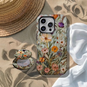 Puede incluir: Una funda de teléfono y un pop socket con un diseño floral de acuarela. La funda es azul claro con flores naranjas, rosas y moradas. Un sombrero de paja y una tela blanca están en el fondo. El pop socket también es floral.