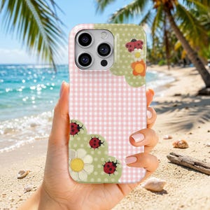 May include: A phone case featuring a pink and white gingham pattern with light green polka dot sections. The design includes illustrations of red ladybugs with black spots and white and orange daisy flowers arranged in the corners.