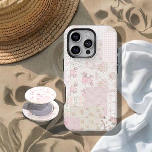 May include: A phone case and pop-up grip with a floral patchwork design. The case features pink and white floral patterns, bows, and gingham. The pop-up grip has a similar floral design. A straw hat and white fabric are in the background.