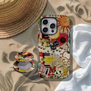 May include: A smartphone case and a matching circular phone grip featuring a collage design with a sun, moon, mushrooms, a frog, apples, musical notes, and the word WHIMSY in cutout letters. The background includes red plaid and sheet music.