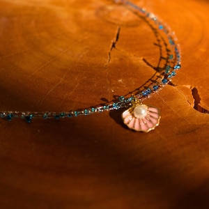 May include: A delicate necklace featuring a double strand of clear and blue beads. A pink seashell pendant with a pearl center hangs from the necklace. The jewelry is set against a warm, wooden background.