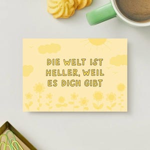 Könnte beinhalten: Eine gelbe Grußkarte mit dem deutschen Text "DIE WELT IST HELLER, WEIL ES DICH GIBT" (Die Welt ist heller, weil es dich gibt). Die Karte zeigt Illustrationen von Sonne, Wolken und Blumen.