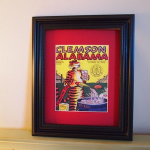 May include: A framed vintage print of a cartoon tiger holding a glass of pink lemonade. The print is yellow, red, and black with the text "Clemson Alabama" and "Pink Lemonade".