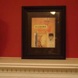 May include: A black framed picture with a vintage sports photo of a football player in a white uniform with the word "ALABAMA" printed on the jersey. The photo is set against a white background with the word "TENNESSEE" printed on the left side of the photo. The photo is framed in a black frame with a white mat.