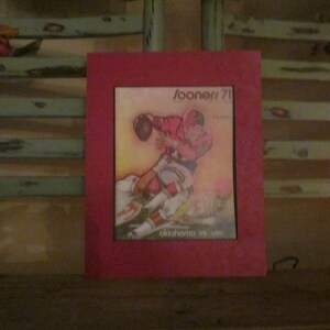 May include: A red framed picture with a vintage print of a football player in action. The print is titled "Sooners '71" and "Oklahoma vs. U.S.C."