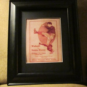 May include: A framed vintage poster advertising a football game between Wabash and Notre Dame on October 13, 1904. The poster features a black and white illustration of a football player in action. The text on the poster reads "Wabash vs. Notre Dame, October 13, 1904, Cartier Field, Price Twenty-Five Cents."