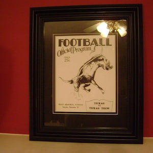 May include: A black framed vintage football program with a drawing of a longhorn bull. The program is for the Texas vs Texas Tech game at Texas Memorial Stadium on September 22. The text reads "Football Official Program Price 25c".