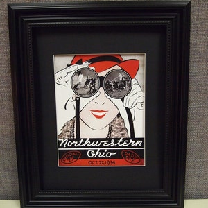 May include: A framed vintage poster advertising a Northwestern Ohio football game on October 27, 1934. The poster features a woman wearing a red hat and looking through binoculars at the game. The text on the poster reads "Northwestern Ohio, Oct. 27, 1934, Official, Dad's Day."