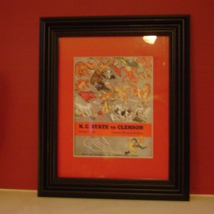 May include: A black framed picture with a red mat and a vintage illustration of a football game between N.C. State and Clemson. The text "N.C. State vs Clemson" is printed in black at the bottom of the illustration.
