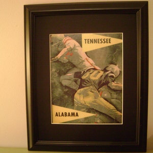 May include: A vintage framed print of a football game between the University of Tennessee and the University of Alabama. The print features two players in action, one in a white jersey with the word "Tennessee" and the other in a green and yellow jersey with the word "Alabama".