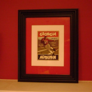 May include: A framed print of a vintage Georgia vs. Auburn football game program from 1951. The program features a black and white photo of a football player in action. The text "Georgia" and "Auburn" are printed in red and white.