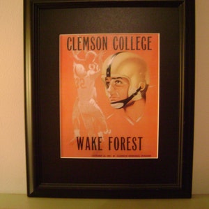 May include: A framed vintage poster advertising a football game between Clemson College and Wake Forest. The poster features a player in a helmet and a red and white background.