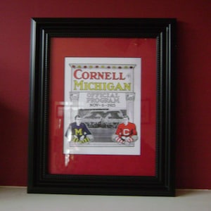 May include: A framed print of a vintage Cornell vs. Michigan hockey game program from November 6, 1915. The program features the text "Cornell Michigan Official Program Nov. 6 - 1915" and two hockey players in their uniforms.