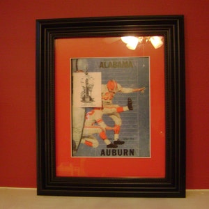 May include: A framed print of a vintage sports illustration featuring two football players in action. The print is in a black frame with a red mat. The text "ALABAMA" and "AUBURN" is visible on the print.