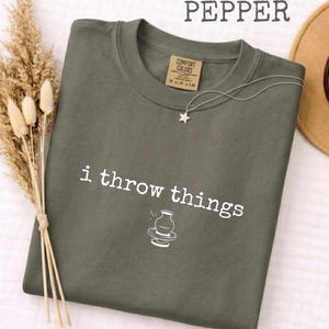 Op de afbeelding: Een donker olijfgroen t-shirt met ronde hals en de witte tekst "i throw things" boven een kleine lijntekening van een pottenbakkersschijf en een vaas. Een zilveren ster-halsketting rust op de halslijn en een beige hoed ligt aan de rechterkant.