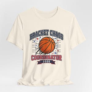 Puede incluir: Camiseta color crema con un gráfico de baloncesto. El diseño incluye el texto "BRACKET CHAOS COORDINATOR 2026" en azul y rojo, con una imagen de baloncesto y elementos decorativos.