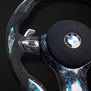 K&ouml;nnte beinhalten: Ein individuelles Autolenkrad mit einem schwarzen, blauen und silbernen Design. Das Lenkrad hat ein Kohlefaser-Muster mit blauen Akzenten, ein schwarzes Mittelpolster mit dem BMW-Logo und integrierte Bedienelemente.