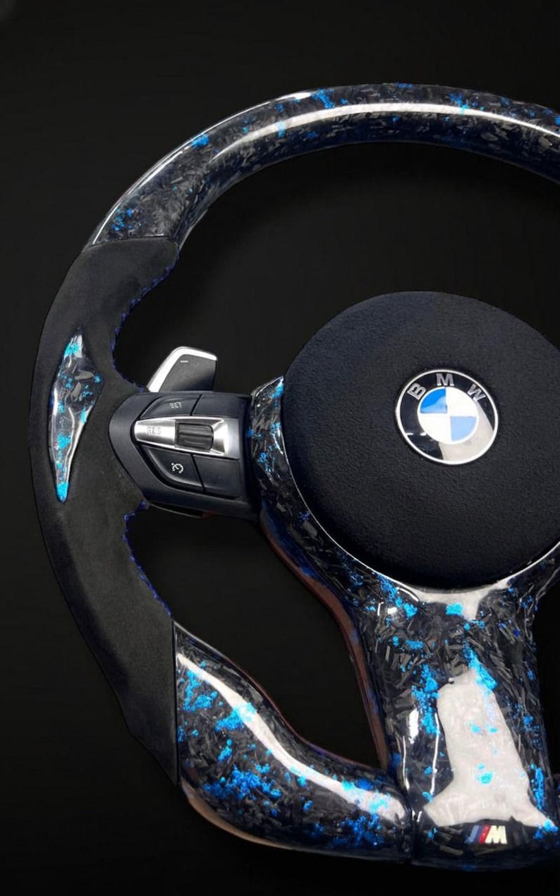 K&ouml;nnte beinhalten: Ein individuelles Autolenkrad mit einem schwarzen, blauen und silbernen Design. Das Lenkrad hat ein Kohlefaser-Muster mit blauen Akzenten, ein schwarzes Mittelpolster mit dem BMW-Logo und integrierte Bedienelemente.