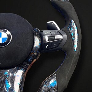 K&ouml;nnte beinhalten: Nahaufnahme eines BMW-Lenkrads mit Carbonfaser- und blauem Akzentdesign. Das Lenkrad verf&uuml;gt &uuml;ber einen schwarzen Wildledergriff, blaue N&auml;hte und integrierte Bedienelemente. Das BMW-Logo ist in der Mitte sichtbar.