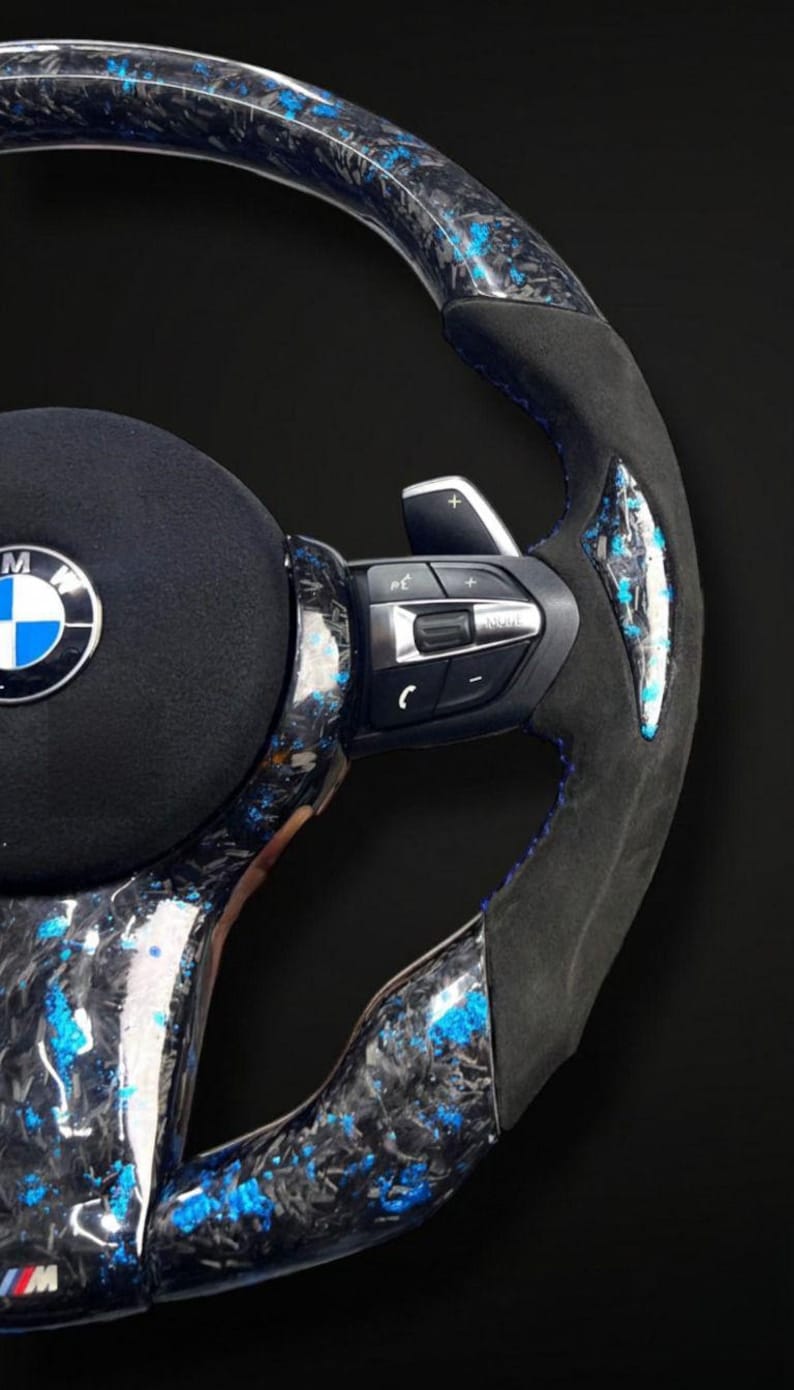 K&ouml;nnte beinhalten: Nahaufnahme eines BMW-Lenkrads mit Carbonfaser- und blauem Akzentdesign. Das Lenkrad verf&uuml;gt &uuml;ber einen schwarzen Wildledergriff, blaue N&auml;hte und integrierte Bedienelemente. Das BMW-Logo ist in der Mitte sichtbar.