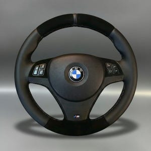 May include: A black and grey BMW steering wheel featuring the BMW emblem at its centre. The wheel has controls on either side, with a suede-like grip at the top and bottom. The M logo is visible at the bottom.