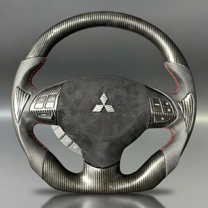 Puede incluir: Un volante de fibra de carbono con costuras rojas. El volante presenta un agarre perforado, botones de control y un centro de gamuza con un emblema plateado. El diseño incorpora una mezcla de texturas y materiales para una estética deportiva.