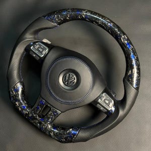 Peut inclure: Un volant en fibre de carbone noir et bleu avec une poign&eacute;e en cuir perfor&eacute;. Le volant pr&eacute;sente des coutures bleues et un logo Volkswagen au centre. La fibre de carbone a un motif mouchet&eacute; bleu et noir.