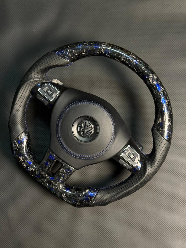 Peut inclure: Un volant en fibre de carbone noir et bleu avec une poign&eacute;e en cuir perfor&eacute;. Le volant pr&eacute;sente des coutures bleues et un logo Volkswagen au centre. La fibre de carbone a un motif mouchet&eacute; bleu et noir.