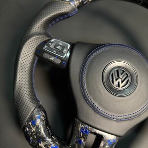 Peut inclure: Un volant de voiture personnalis&eacute; avec une poign&eacute;e en cuir noir et des coutures bleues. Le volant pr&eacute;sente un design en fibre de carbone avec des accents bleus et un logo Volkswagen au centre. Le volant poss&egrave;de des boutons de commande int&eacute;gr&eacute;s.