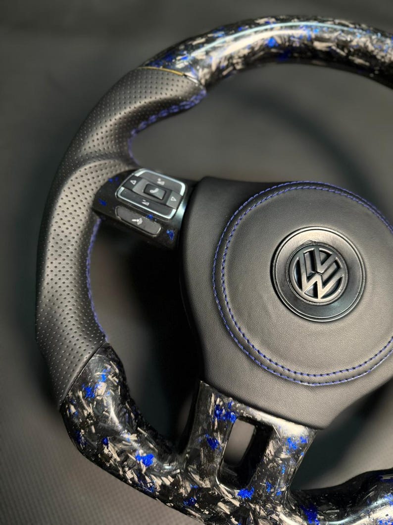 Peut inclure: Un volant de voiture personnalis&eacute; avec une poign&eacute;e en cuir noir et des coutures bleues. Le volant pr&eacute;sente un design en fibre de carbone avec des accents bleus et un logo Volkswagen au centre. Le volant poss&egrave;de des boutons de commande int&eacute;gr&eacute;s.
