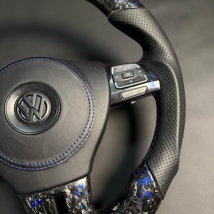 Peut inclure: Gros plan sur un volant noir et bleu avec le logo Volkswagen. Le volant pr&eacute;sente un design en fibre de carbone avec des accents bleus et des poign&eacute;es en cuir perfor&eacute;. Des coutures bleues sont visibles sur la partie centrale.