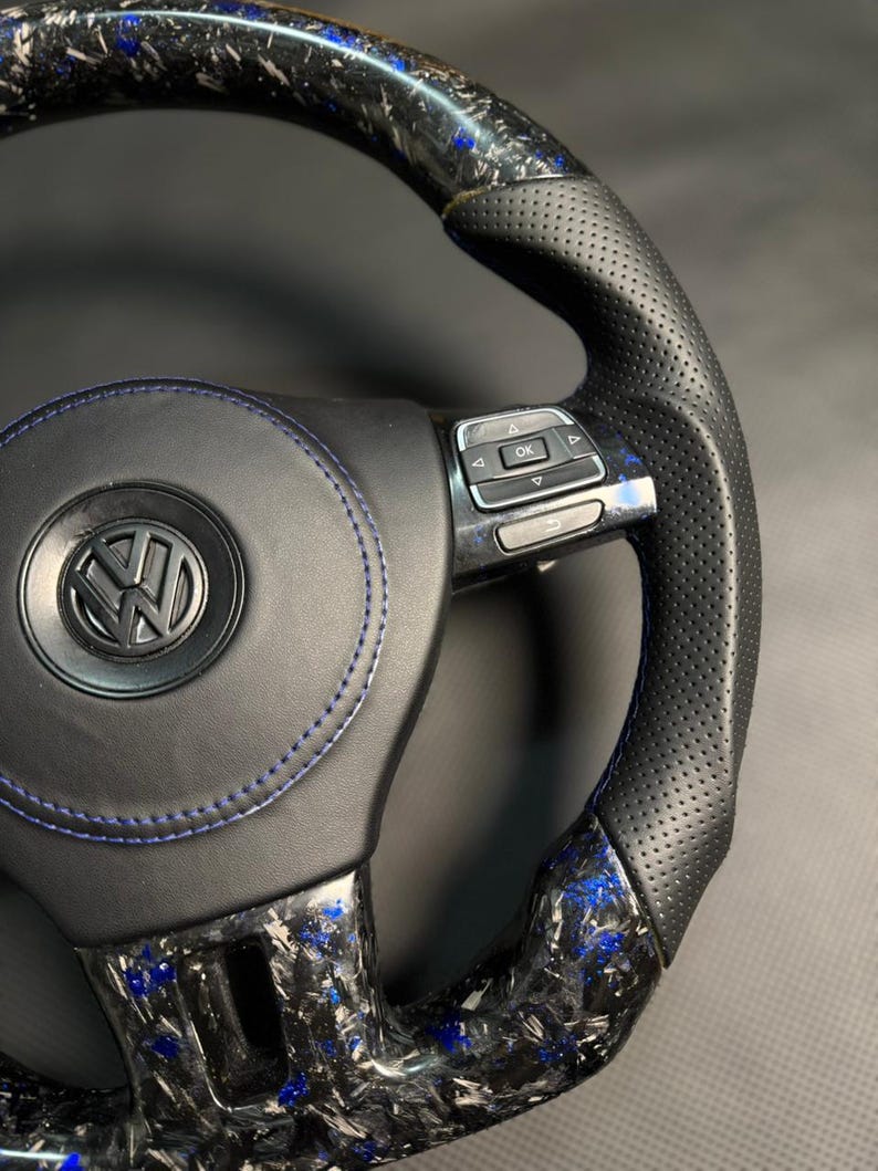 Peut inclure: Gros plan sur un volant noir et bleu avec le logo Volkswagen. Le volant pr&eacute;sente un design en fibre de carbone avec des accents bleus et des poign&eacute;es en cuir perfor&eacute;. Des coutures bleues sont visibles sur la partie centrale.