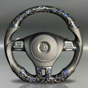 Peut inclure: Un volant de voiture personnalis&eacute; avec une poign&eacute;e en cuir noir et un design avec des accents en fibre de carbone et bleu. Le centre pr&eacute;sente un embl&egrave;me noir avec le logo Volkswagen. Le volant a des boutons de commande int&eacute;gr&eacute;s.
