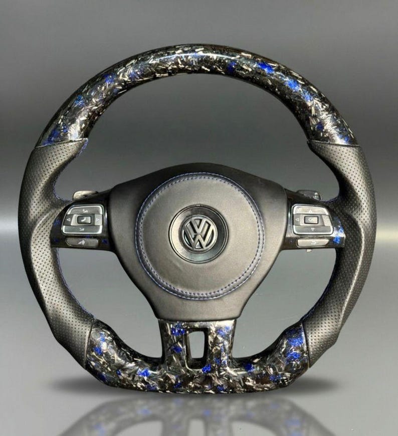 Peut inclure: Un volant de voiture personnalis&eacute; avec une poign&eacute;e en cuir noir et un design avec des accents en fibre de carbone et bleu. Le centre pr&eacute;sente un embl&egrave;me noir avec le logo Volkswagen. Le volant a des boutons de commande int&eacute;gr&eacute;s.
