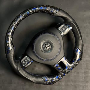 Peut inclure: Un volant en fibre de carbone noir et bleu avec une poign&eacute;e en cuir perfor&eacute;. Le centre pr&eacute;sente un embl&egrave;me noir avec un logo argent&eacute;. Le volant poss&egrave;de des boutons de commande int&eacute;gr&eacute;s et des coutures bleues.