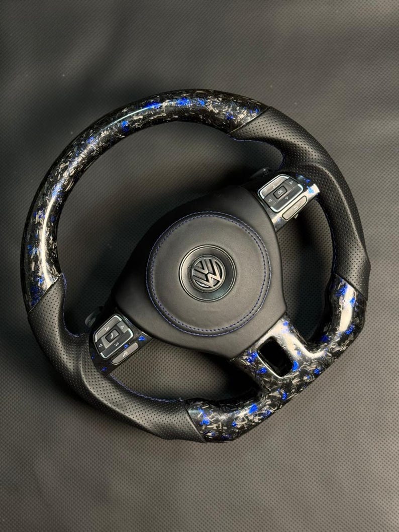 Peut inclure: Un volant en fibre de carbone noir et bleu avec une poign&eacute;e en cuir perfor&eacute;. Le centre pr&eacute;sente un embl&egrave;me noir avec un logo argent&eacute;. Le volant poss&egrave;de des boutons de commande int&eacute;gr&eacute;s et des coutures bleues.