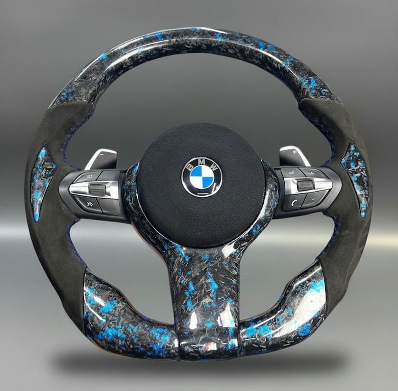 K&ouml;nnte beinhalten: Ein individuelles Lenkrad mit einem Carbonfaser- und blauen Design. Das Lenkrad hat einen schwarzen Wildledergriff und Schaltwippen. In der Mitte des Lenkrads befindet sich das BMW-Logo.