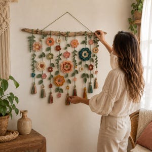 Op de afbeelding: Een handgemaakte wandhanger met een houten tak met gehaakte bloemen in verschillende kleuren, waaronder perzik, roze en blauwgroen. Groene bladeren en kwasten dragen bij aan het decoratieve ontwerp. De wandhanger wordt op een witte muur getoond.