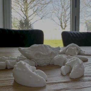 May include: A white 3D-printed crab sculpture with detailed claws and body segments. The crab is positioned on a wooden surface, with a blurred background of a window and trees. The sculpture is a decorative item.