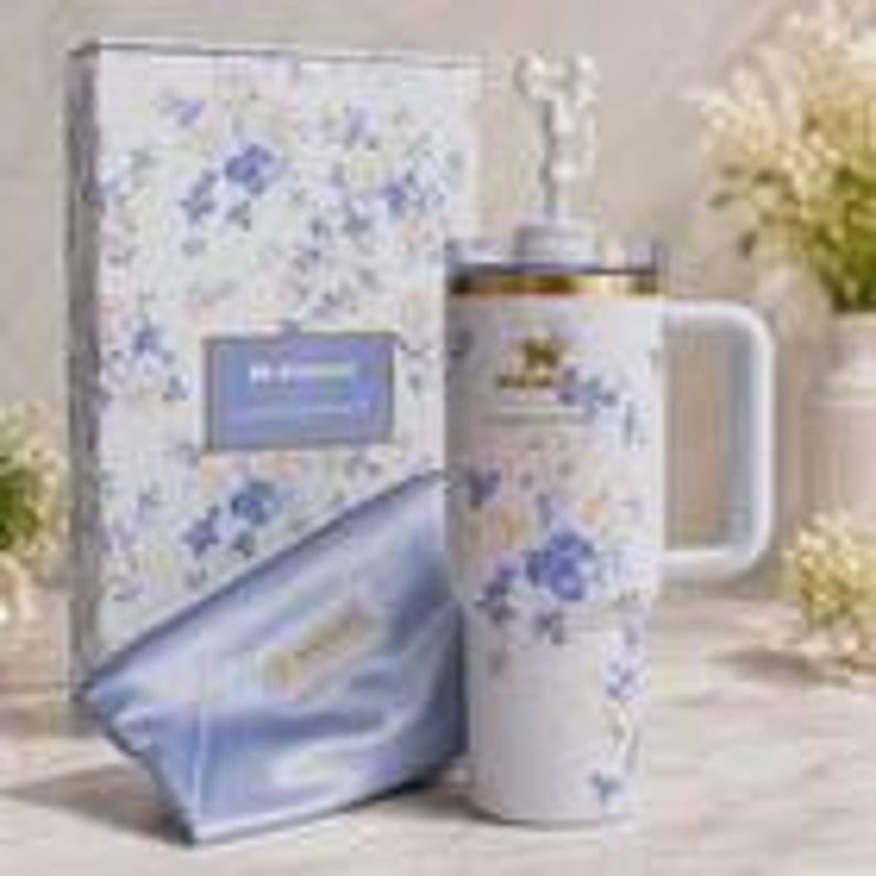 May include: A white Stanley tumbler with a floral design, a handle, and a lid with a straw. The tumbler is next to a matching gift box and a satin pouch. The floral pattern includes blue and yellow flowers on a white background.