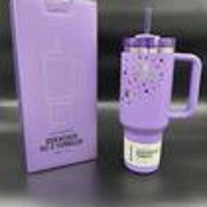 Puede incluir: Un vaso t&eacute;rmico lavanda con asa y pajita, junto a una caja a juego. El vaso presenta una tapa plateada y un dise&ntilde;o de corazones y estrellas moradas. La caja tiene texto y un gr&aacute;fico del vaso.