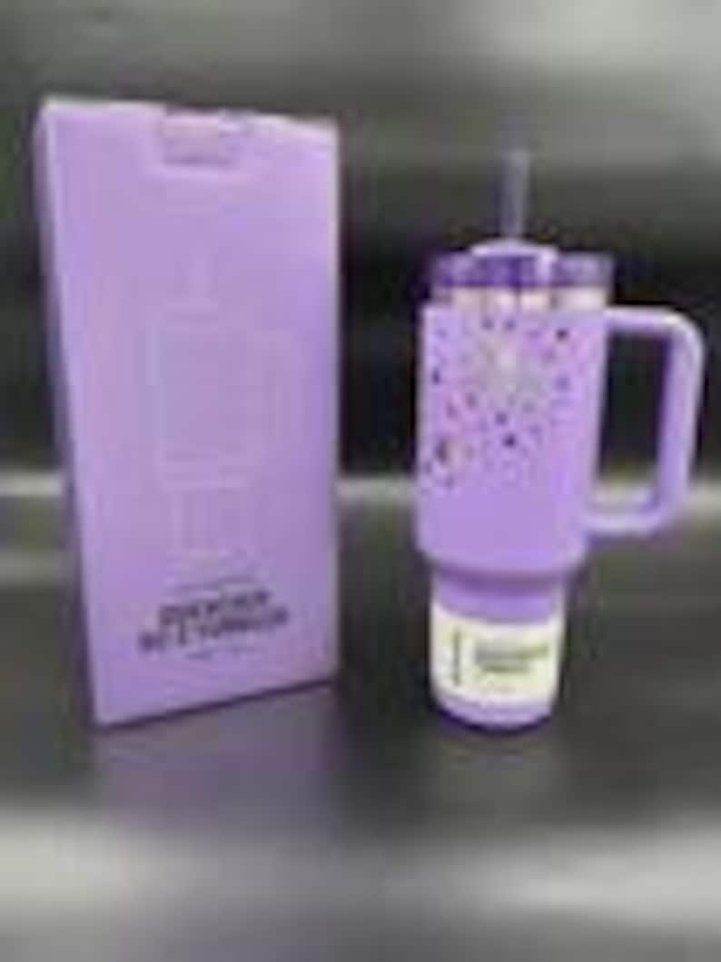 Puede incluir: Un vaso t&eacute;rmico lavanda con asa y pajita, junto a una caja a juego. El vaso presenta una tapa plateada y un dise&ntilde;o de corazones y estrellas moradas. La caja tiene texto y un gr&aacute;fico del vaso.